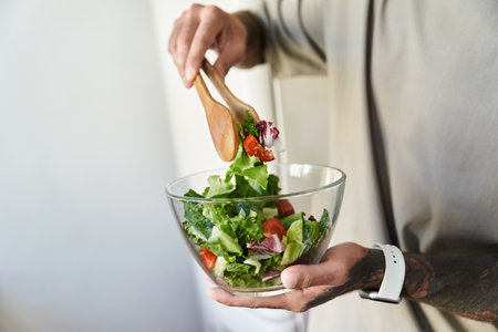 This young man skillfully tosses a vibrant salad in a glass bowl, blending fresh ingredients.の写真素材