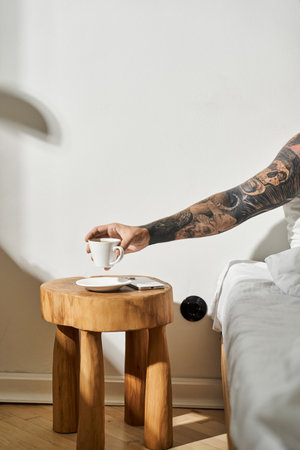 A young man at home taking his coffee while relaxing in bed by a unique wooden table.
