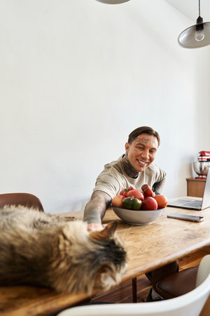A cheerful young man reaches out to his cat while surrounded by a bowl of fresh fruit at home.の写真素材
