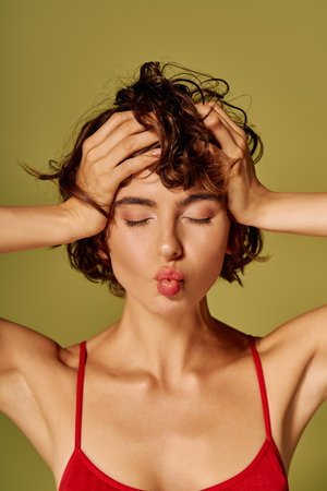 A young woman holds her head, showing a mix of emotions while playfully pouting her lips.