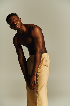 A young handsome African American man stands confidently, posing topless while wearing tailored beige pants. His posture exudes elegance and self assurance, set against a minimalist backdrop.の写真素材