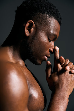 A young handsome African American man poses topless with a thoughtful expression. His hands are clasped in prayer as he gazes downwards, evoking a sense of tranquility and introspection.の写真素材