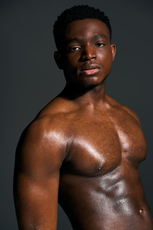 A young African American man stands confidently, showcasing his sculpted torso against a dark backdrop. His expression is captivating, reflecting strength and charisma in this striking portrait.の写真素材