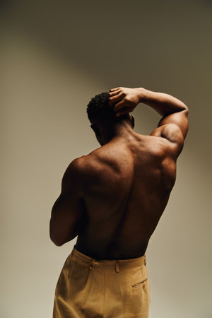 A confident young man stands with a strong posture, his back facing the viewer. He radiates charisma and style, highlighted by soft, artistic lighting that enhances his muscular form.の写真素材