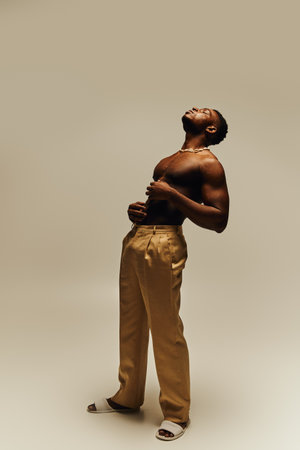 This striking portrait features a young, confident black man posing topless with a relaxed expression. His stylish beige pants complement his athletic build, creating an impressive visual contrast.の写真素材