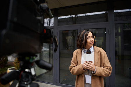 A journalist in a beige coat holds a microphone, thoughtfully preparing to report news outside a...