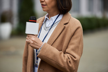 A poised multiracial woman holds a microphone, ready to deliver breaking news on a lively city street.の写真素材