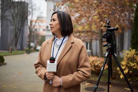 A multiracial journalist wearing a beige coat stands outdoors with a microphone, preparing to share news updates.の写真素材