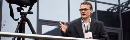 A focused journalist in a coat speaks into a microphone outside a contemporary structure during...
