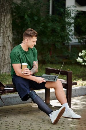 A young man with a prosthetic leg relaxes on a bench, sipping coffee and focused on his laptop in a...