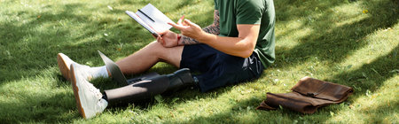 A young man with a prosthetic leg sits on grass, absorbed in reading a book, embracing nature and...