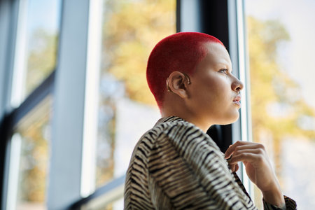 A bald woman with striking red hair looks pensively outside, reflecting in the warm sunlight.の写真素材