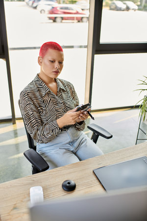 A bald woman with vibrant red hair intently uses her phone while seated at a stylish desk.の写真素材