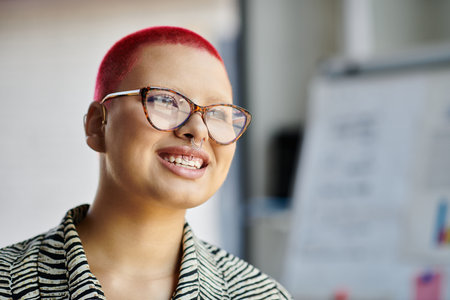 A confident bald woman sporting a stylish red haircut and glasses smiles warmly in a bright room.の写真素材