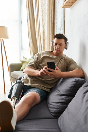 Handsome young man sits comfortably on a couch, focused on his smartphone, wearing headphones.の写真素材