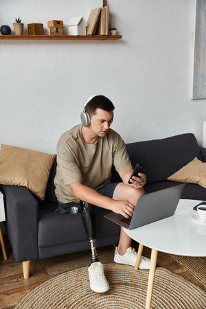Young handsome man with prosthetic leg engages with laptop and smartphone in living room.の写真素材