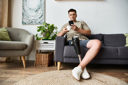 Young man sits comfortably on a couch, enjoying a drink and looking at his phone.の写真素材