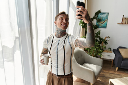 Handsome young man with tattoos smiles while taking a selfie and holding a cup of coffee.の写真素材