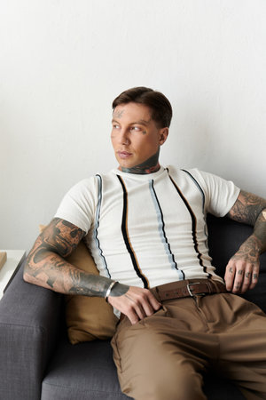 Young man with tattoos sits casually on a modern couch in a chic apartment.の写真素材