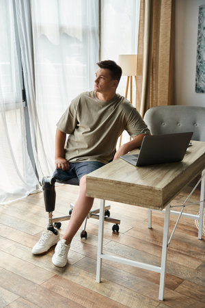 Handsome young man sits in a stylish room, focused on a laptop at a wooden desk.の写真素材