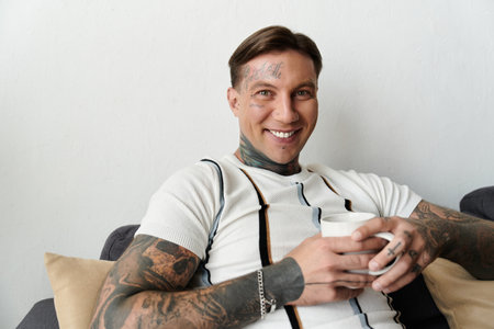 Smiling young man with tattoos enjoys a cup while sitting on a couch in a bright room.の写真素材
