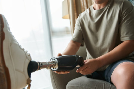 Young handsome man sits comfortably while adjusting his prosthetic leg in a cozy setting.の写真素材