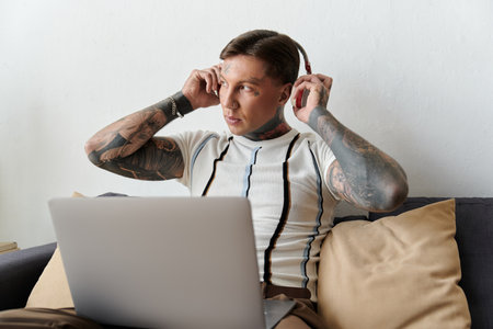 Young man with tattoos listens to music on headphones while using a laptop at home.の写真素材