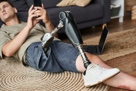 Young man comfortably sitting on a rug, focused on his smartphone, showcasing his prosthetic leg.の写真素材