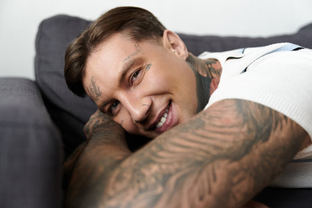 Young man with tattoos relaxes on a couch, showcasing a bright smile and engaging demeanor.の写真素材