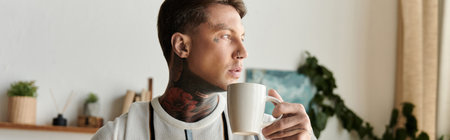A confident young man with tattoos enjoys a warm cup of coffee while relaxing indoors.の写真素材
