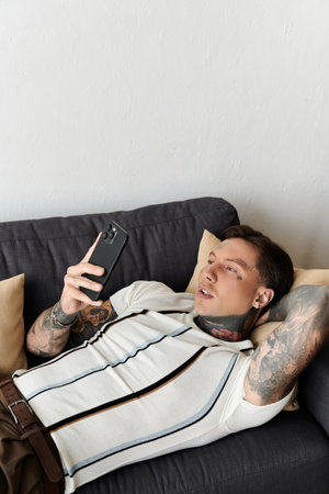 Handsome young man with tattoos lounging comfortably on a couch looking at his smartphone.の写真素材
