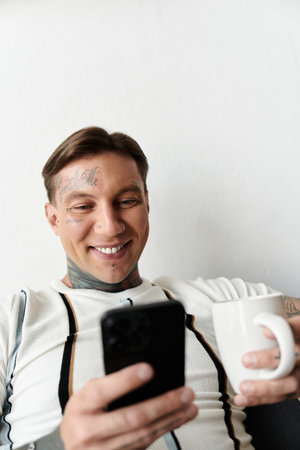Man with tattoos smiles while sitting comfortably and checking his phone with a coffee mug.の写真素材