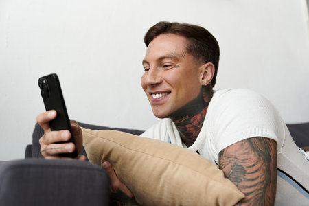 Young man with tattoos relaxes on a couch, enjoying time on his smartphone and smiling.の写真素材
