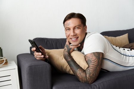 Man with tattoos enjoys a moment of relaxation while using his smartphone on a couch.の写真素材