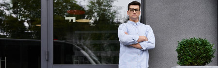 Handsome young man poses with arms crossed near a contemporary structure in daylight.の写真素材
