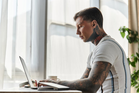 Handsome young man with tattoos focuses on his laptop in a stylish, well lit room.の写真素材