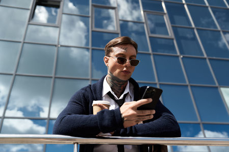 Man with tattoos enjoys coffee while looking at his smartphone near a glass structure.の写真素材