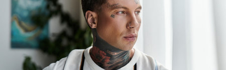 Young man with tattoos gazes thoughtfully while standing near a window with natural light.の写真素材