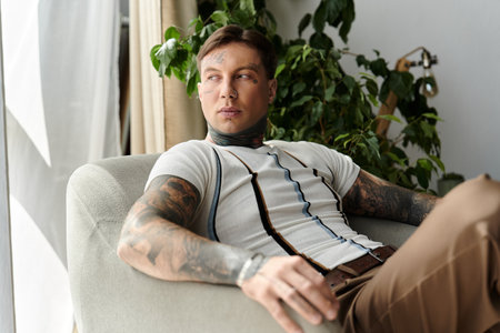 A young handsome man with tattoos is sitting comfortably in a modern room surrounded by plants.の写真素材