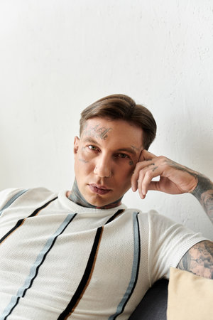 A young man with striking tattoos relaxes comfortably on a sofa in a modern room.の写真素材