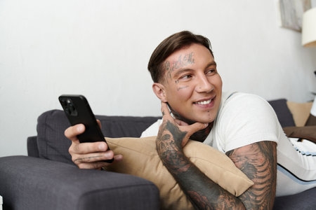 Handsome man lounging on a couch with tattoos, casually checking his smartphone and smiling.の写真素材