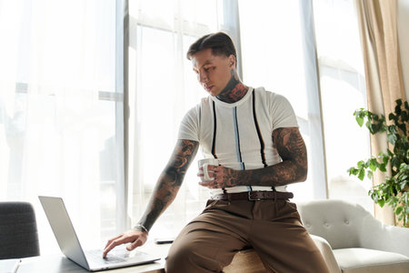 Young man with tattoos sits on a table using a laptop while holding a cup in a stylish room.の写真素材