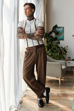 Handsome young man with tattoos poses by a window, dressed in stylish attire with a relaxed vibe.の写真素材