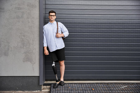 A young man with a prosthetic leg leans casually against a garage wall while wearing stylish attire.