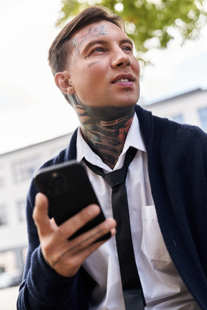 Handsome young man with tattoos sits casually, using his phone while enjoying the sunshine.の写真素材