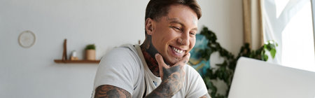 A cheerful young man with tattoos enjoys his time at a laptop in a cozy indoor space.の写真素材