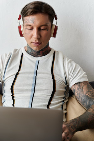 A young man with tattoos enjoys music through headphones as he works on his laptop at home.の写真素材