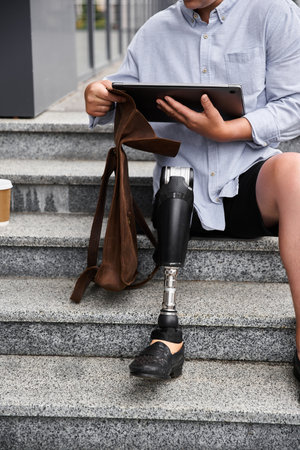 A young man with a prosthetic leg sits on stone stairs while using a tablet and holding a bag.