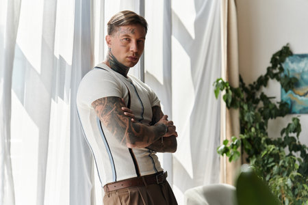 Young man with tattoos poses confidently, showcasing his style in a sunlit room.