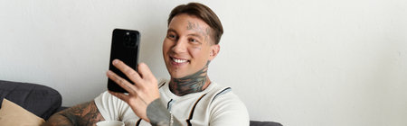 Handsome young man with tattoos smiles while holding his smartphone and sitting comfortably.の写真素材